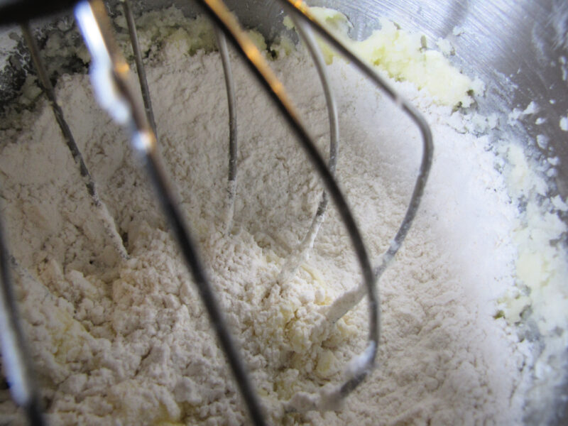 Adding flour mixture to butter and sugar while making cherry almond chocolate chip cookie dough in a stand mixer. | Gathered In The Kitchen 