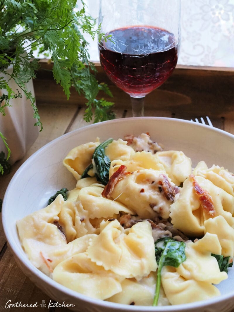 A white ceramic serving bowl filled with cheesy cream sauce tortellini, fresh green baby spinach leaves, and crispy bacon pieces next to a glass of red wine. | Gathered In The Kitchen 