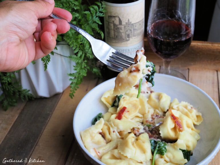 A hand holding a silver fork lifting a cheesy piece of pasta with wilted spinach and crispy bacon out of a white bowl. | Gathered In The Kitchen