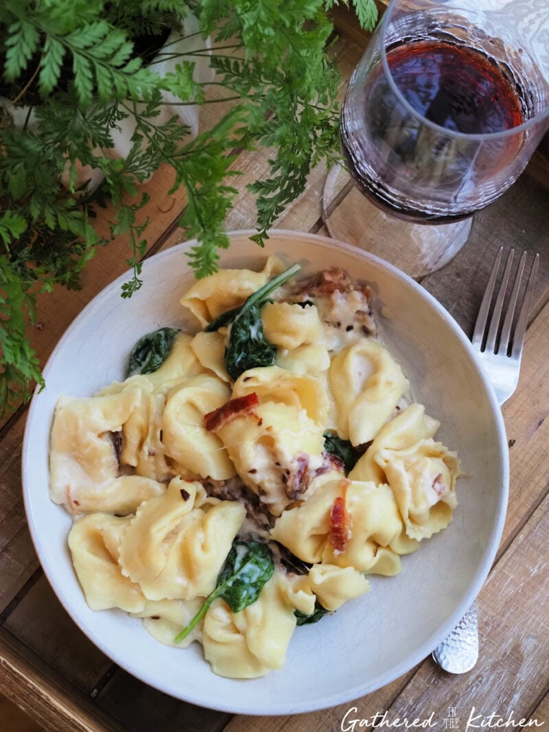 An overhead view of a bowl of creamy cheese tortellini with green spinach and red bacon bits sitting on a rustic wooden tray next to a wine glass and fork. | Gathered In The Kitchen