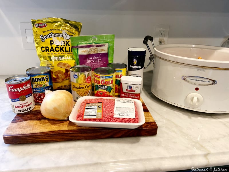 Ingredients for BBQ Pork Rind Cheesy Casserole including ground beef, kidney beans, corn, diced tomatoes, onion, cheese, cream of mushroom soup, pork rinds, and spices next to a slow cooker.