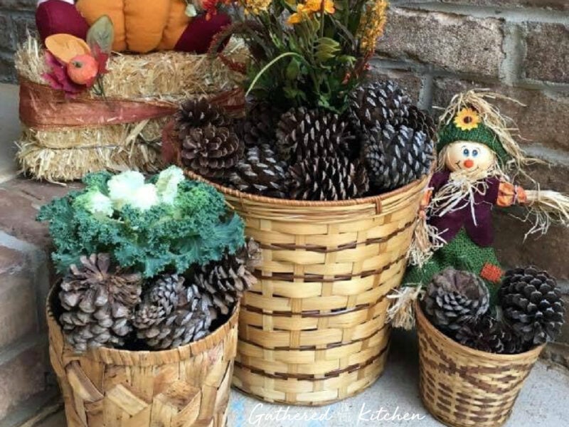 A fall porch display featuring wicker baskets filled with pinecones, ornamental kale, a small scarecrow, and a hay bale with decorative pumpkins, creating a cozy autumn vibe. | Gathered In The Kitchen