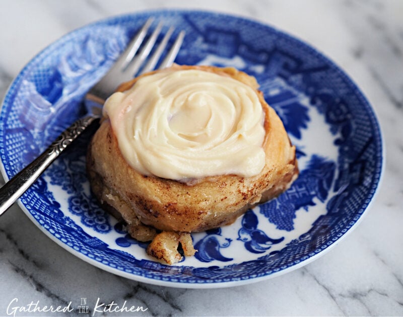 A warm, homemade cinnamon roll topped with creamy 3-ingredient cinnamon roll frosting made with cream cheese and maple syrup, served on a blue and white vintage plate with a silver fork. Perfect for breakfast or a cozy weekend brunch.