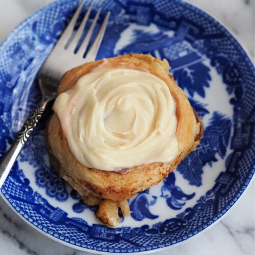 A warm homemade cinnamon roll topped with creamy 3-ingredient frosting, served on a blue and white vintage plate with a silver fork. The cream cheese and maple syrup frosting is smooth, thick, and ready to melt into the soft layers of the roll. Perfect for a quick and easy cinnamon roll glaze.