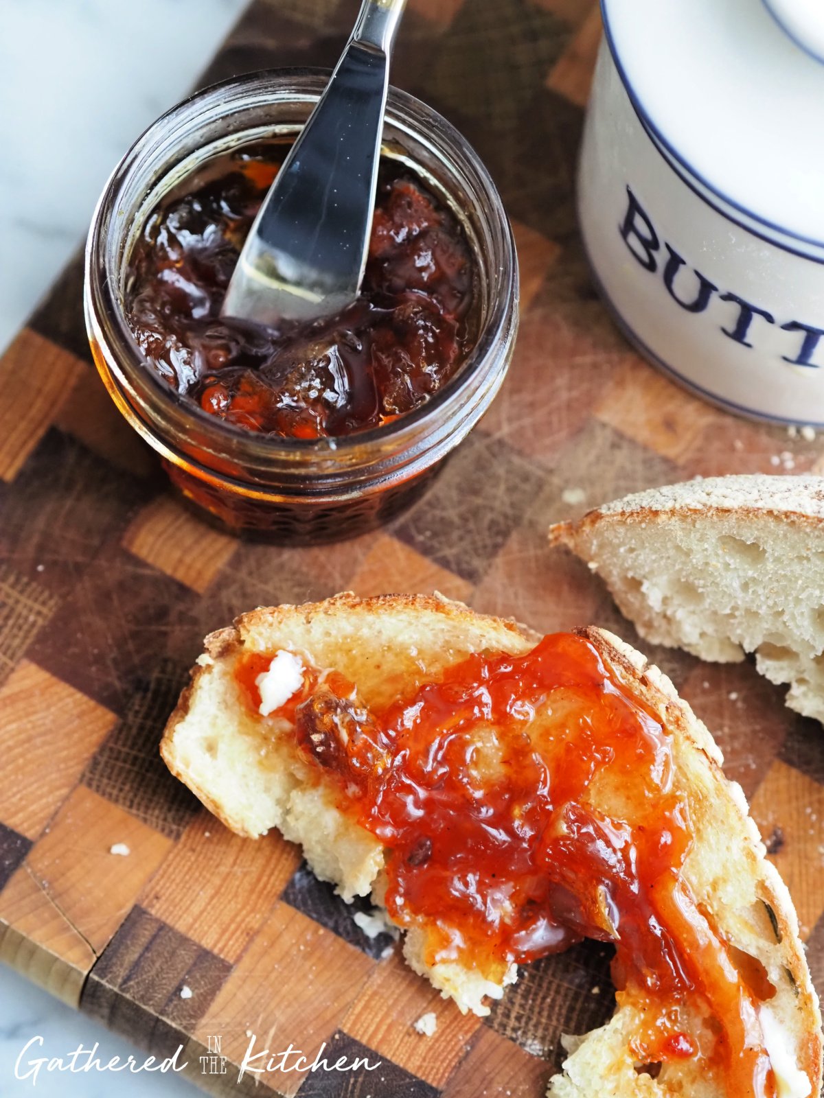 A jar of homemade peach conserve with rum sits on a rustic wooden cutting board, with a butter knife dipped in the rich, chunky spread. A slice of fresh sourdough bread is topped with the glistening conserve, paired with a butter dish in the background. The vibrant orange and red hues of the preserve showcase the fresh fruit and warm spices inside | Gathered In The Kitchen