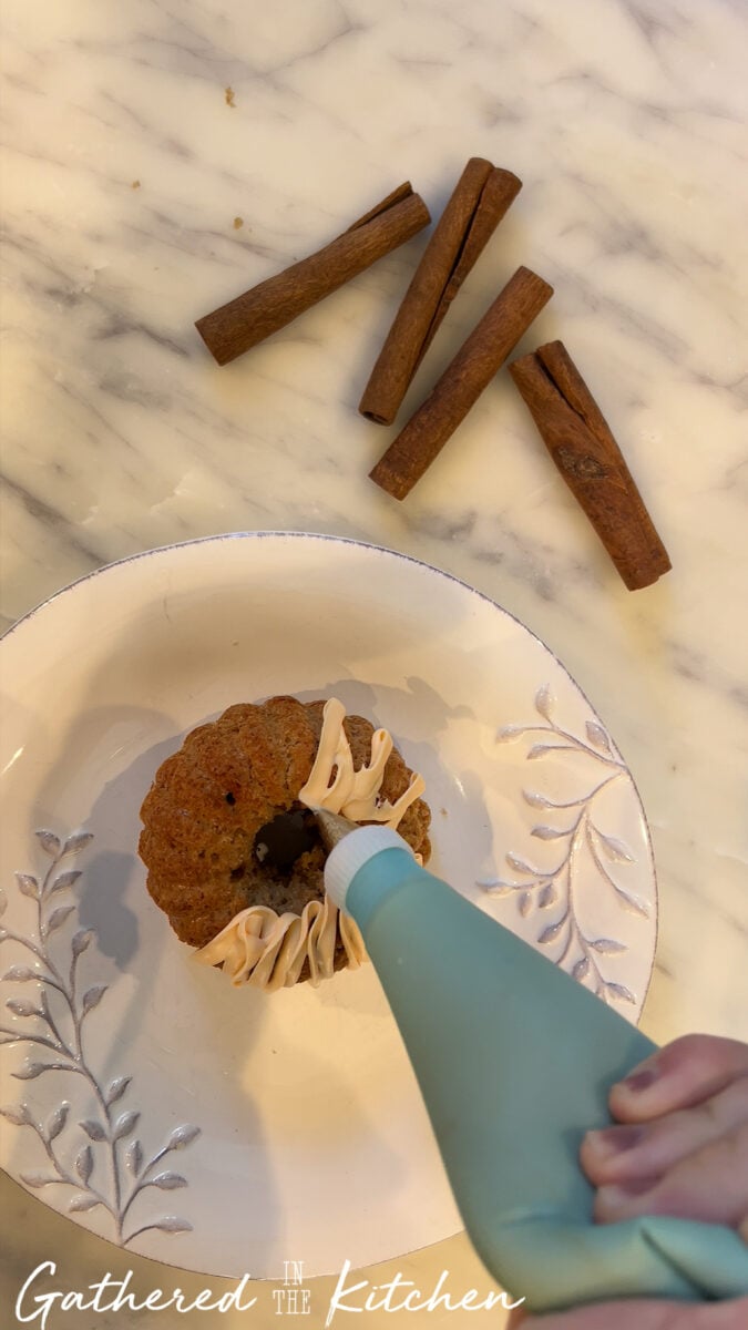 Piping cream cheese frosting onto a mini bundt cake before assembling into pumpkin-shaped cakes with cinnamon stick stems. | Gathered In The Kitchen 