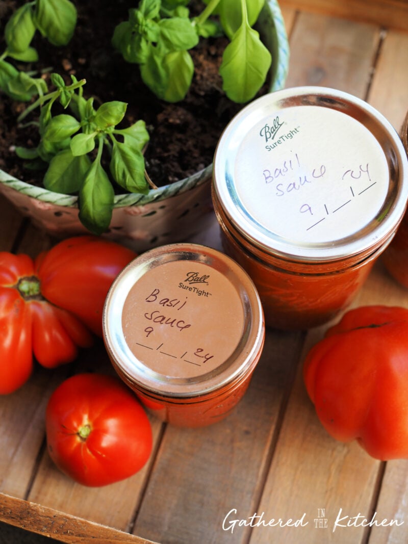 Two jars of homemade tomato basil sauce labeled with the canning date, sitting beside fresh tomatoes and basil plants, showing the finished result of water bath canning. | Gathered In The Kitchen 