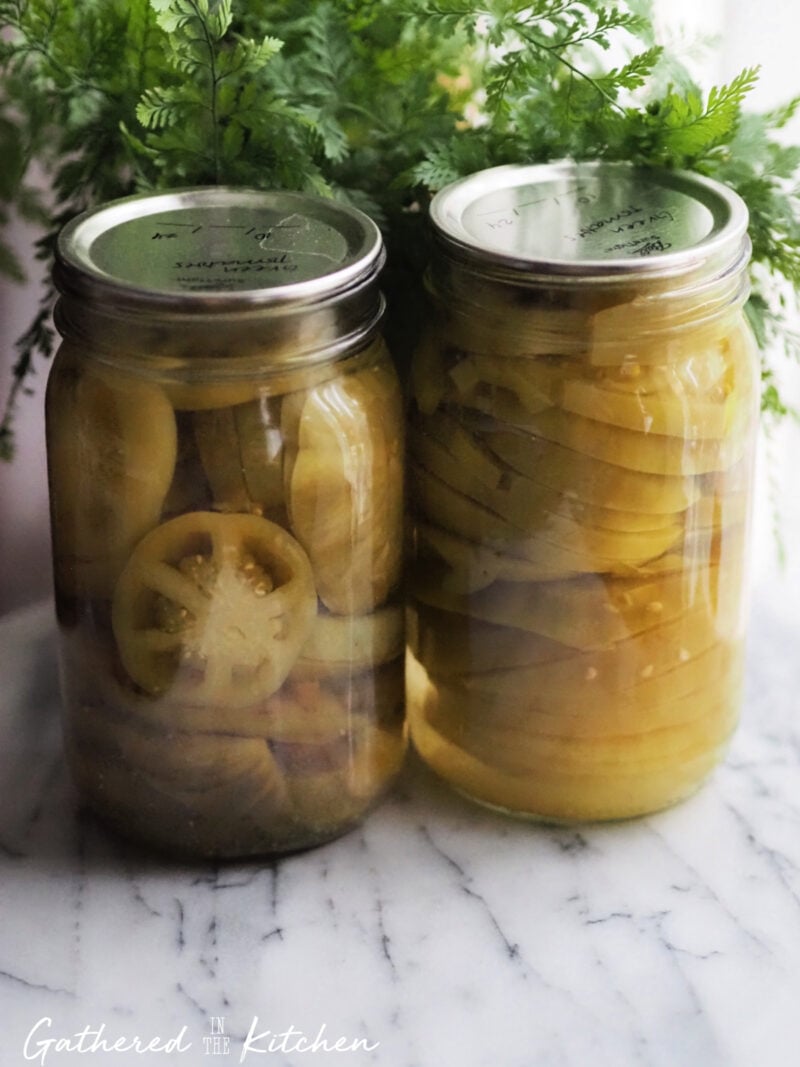 Two glass jars filled with sliced green tomatoes preserved in liquid after water bath canning, sitting on a marble counter with fresh greenery in the background. | Gathered In The Kitchen 