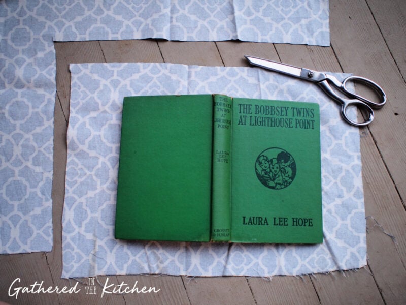 Green vintage hardback book placed on top of cut blue patterned cotton fabric with scissors, showing how to size and trim material for a DIY fabric book cover project.