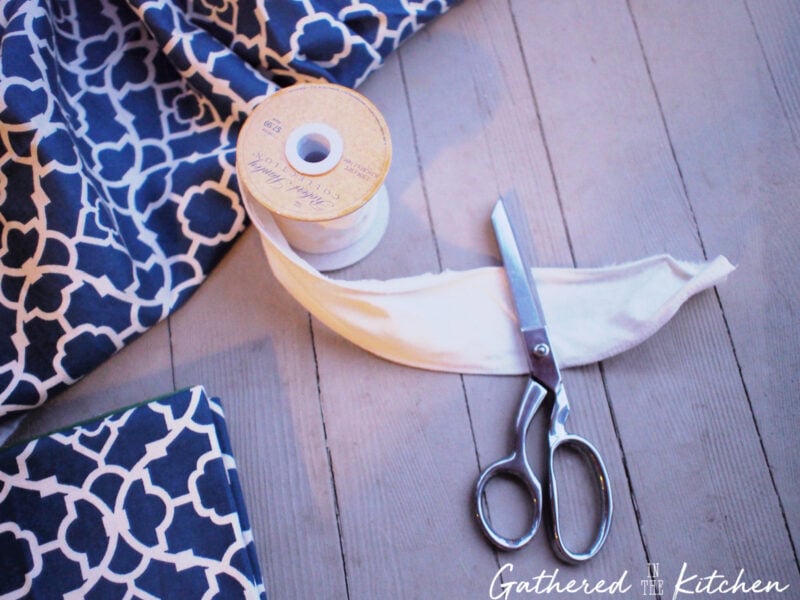 Scissors, cream-colored ribbon, and blue patterned cotton fabric laid on wood surface, showing supplies for finishing and styling a DIY no-sew fabric book cover.