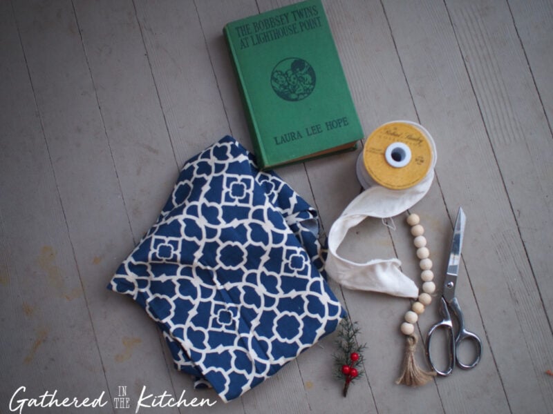 Flat lay of DIY fabric book cover supplies including a green hardback book, blue patterned cotton fabric, scissors, ribbon, wood beads, greenery, and hot glue gun materials—perfect for making a handmade cloth book jacket.