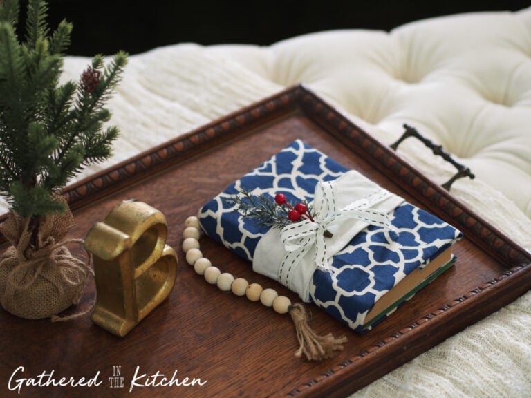 DIY no-sew fabric book cover made with blue patterned cloth, tied with ribbon and greenery, styled on a wooden tray with a Christmas tree, wooden beads, and holiday decorations.