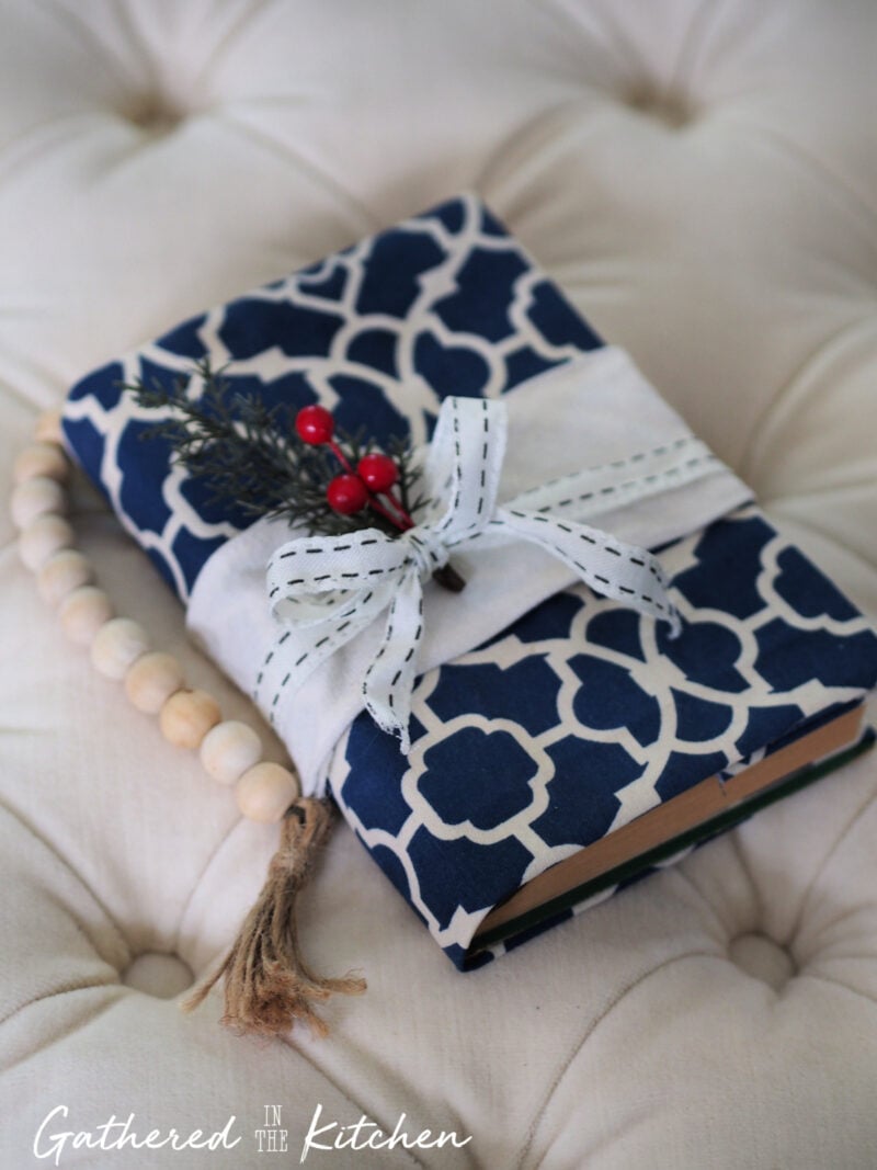 Blue patterned DIY fabric book cover tied with a stitched white ribbon and decorated with greenery and red berries, styled with a wooden bead garland—perfect as a no-sew handmade gift idea.