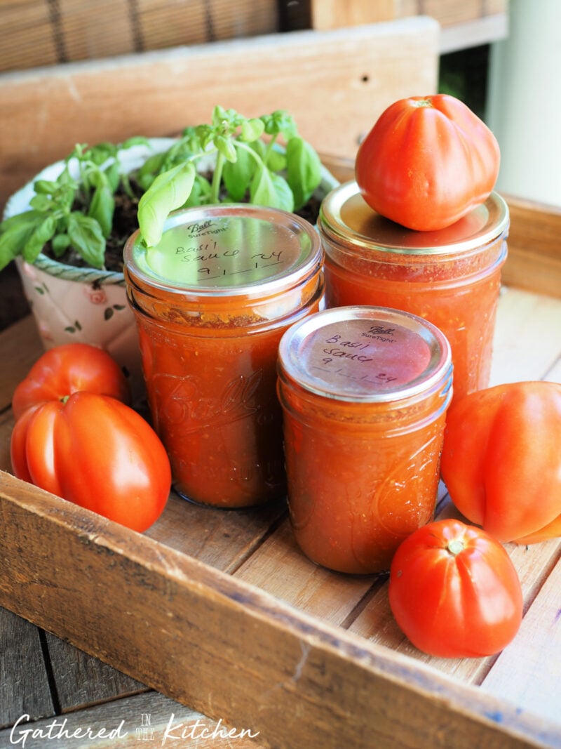Three jars of homemade tomato basil sauce with handwritten labels, displayed in a wooden crate alongside fresh tomatoes and basil plants — the finished result of water bath canning. | Gathered In The Kitchen 