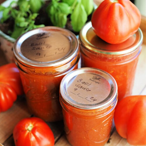 Jars of homemade tomato basil sauce preserved with water bath canning, sitting on a wooden tray with fresh garden tomatoes. | Gathered In The Kitchen