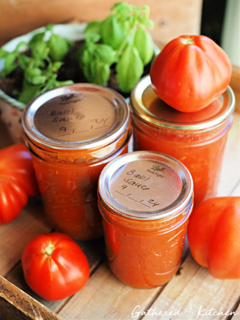 Jars of homemade tomato basil sauce preserved with water bath canning, sitting on a wooden tray with fresh garden tomatoes. | Gathered In The Kitchen