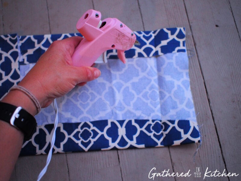 Hand holding a pink hot glue gun, applying glue to the folded edges of blue patterned cotton fabric to create pockets for a DIY no-sew fabric book cover.