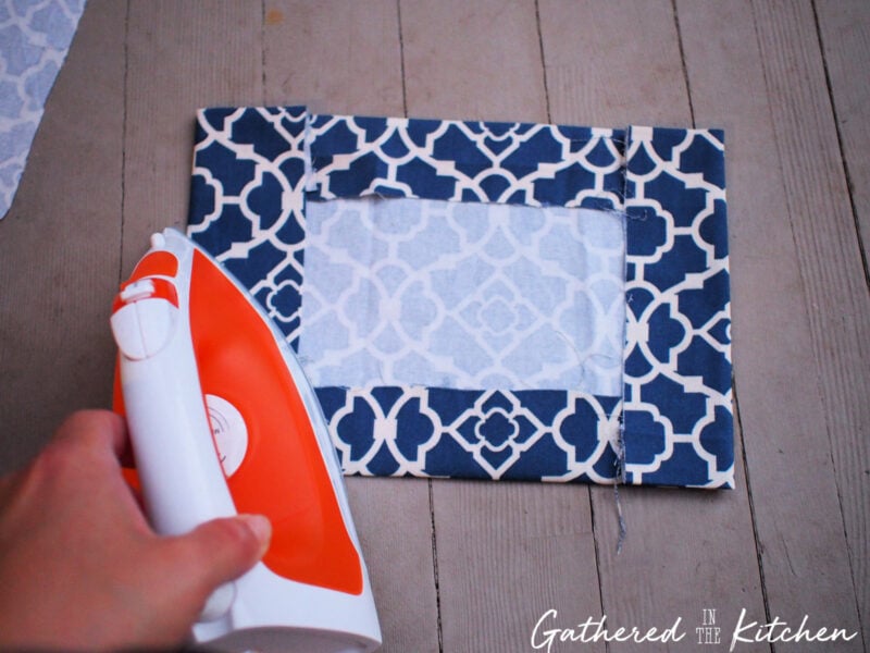 Hand holding an orange and white iron, pressing folded blue patterned cotton fabric edges to prepare crisp hems for a DIY no-sew fabric book cover tutorial.