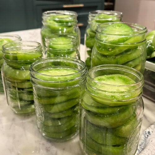 Several glass jars filled with neatly packed slices of fresh green tomatoes on a kitchen counter, prepared for water bath canning. | Gathered In The Kitchen