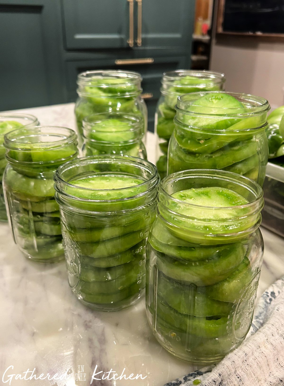 Several glass jars filled with neatly packed slices of fresh green tomatoes on a kitchen counter, prepared for water bath canning. | Gathered In The Kitchen