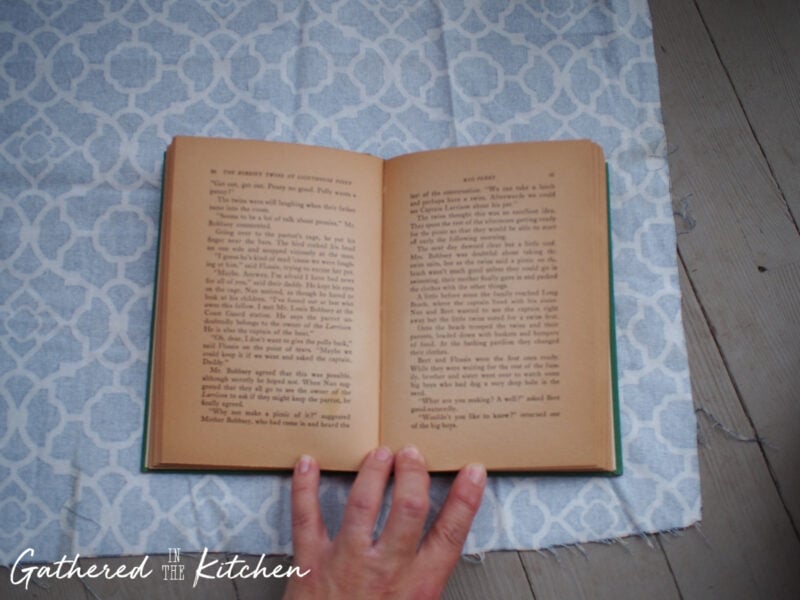 Open vintage hardback book laid on patterned blue cotton fabric, showing how to measure and cut material for a DIY fabric book cover tutorial.