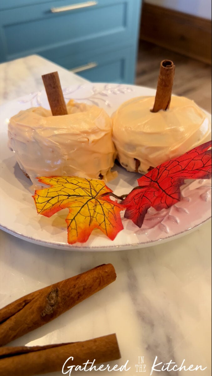 Frosted pumpkin-shaped mini bundt cakes decorated with orange icing and cinnamon stick stems on a fall plate. | Gathered In The Kitchen 