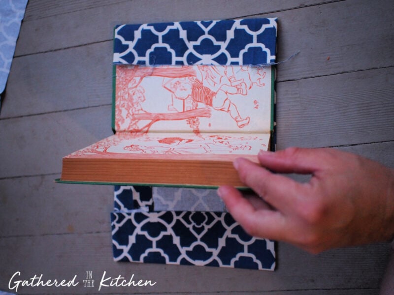 Hand sliding a vintage hardback book into blue patterned fabric pockets to finish a DIY no-sew fabric book cover.