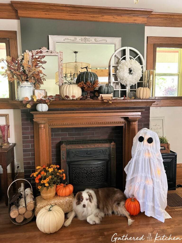 DIY Ghost Christmas Tree made from an artificial prelit tree draped with a white ruffled curtain and black felt eyes, styled next to pumpkins, flowers, and a decorated fall mantle with a dog resting in front. | Gathered In The Kitchen
