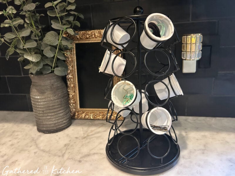A black Keurig coffee pod holder repurposed into a spinning jewelry and hair accessory organizer, with empty white K Cups holding earrings, bobby pins, and hair ties. Displayed on a marble countertop with a vase and gold frame in the background. | Gathered In The Kitchen 