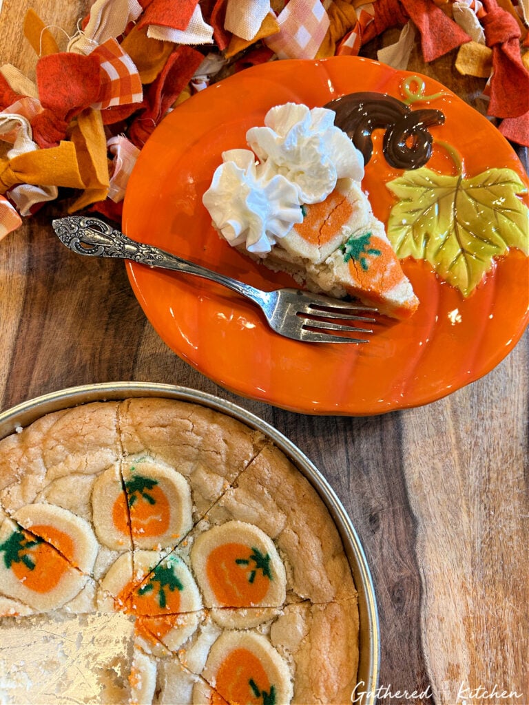 Slice of Pillsbury pumpkin sugar cookie cake served on an orange fall plate with whipped cream, next to the full cookie pie baked in a round cake pan. | Gathered In The Kitchen