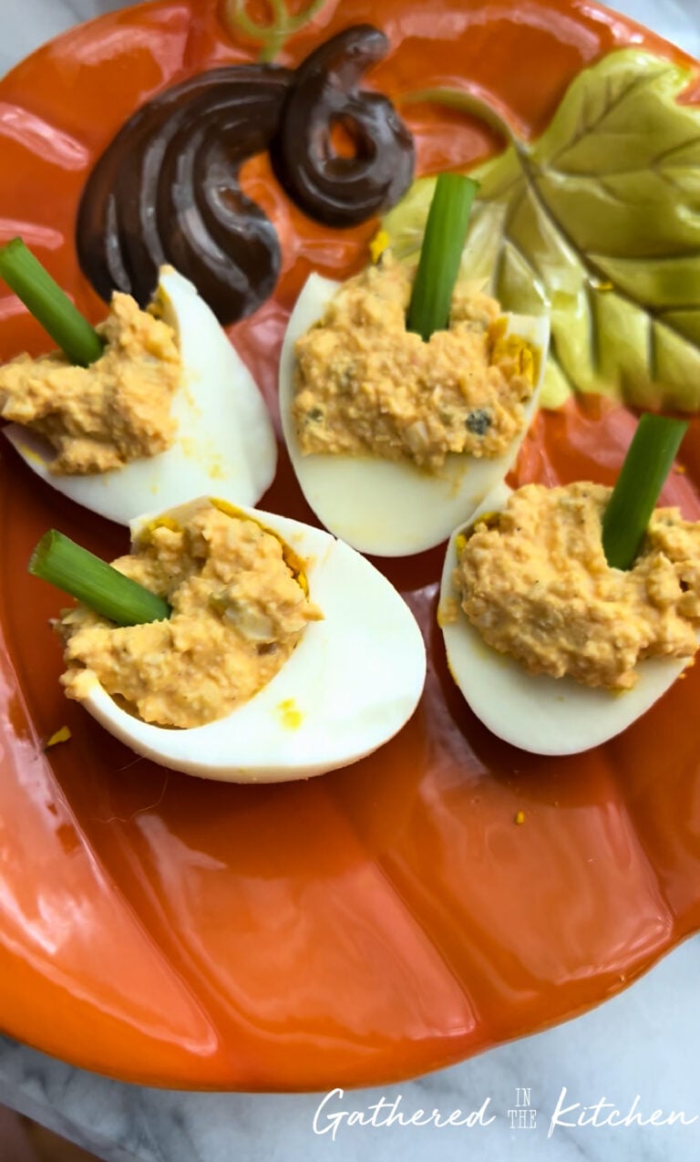 Four deviled eggs styled to look like pumpkins with green onion stems on a pumpkin-shaped fall plate. | Gathered In The Kitchen