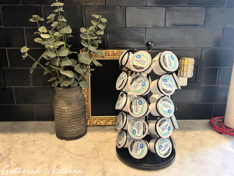Original Keurig K Cup holder filled with donut shop coffee pods, sitting on a marble kitchen counter next to a gray vase and a gold picture frame. Ready for a creative repurposing DIY project. | Gathered In The Kitchen 