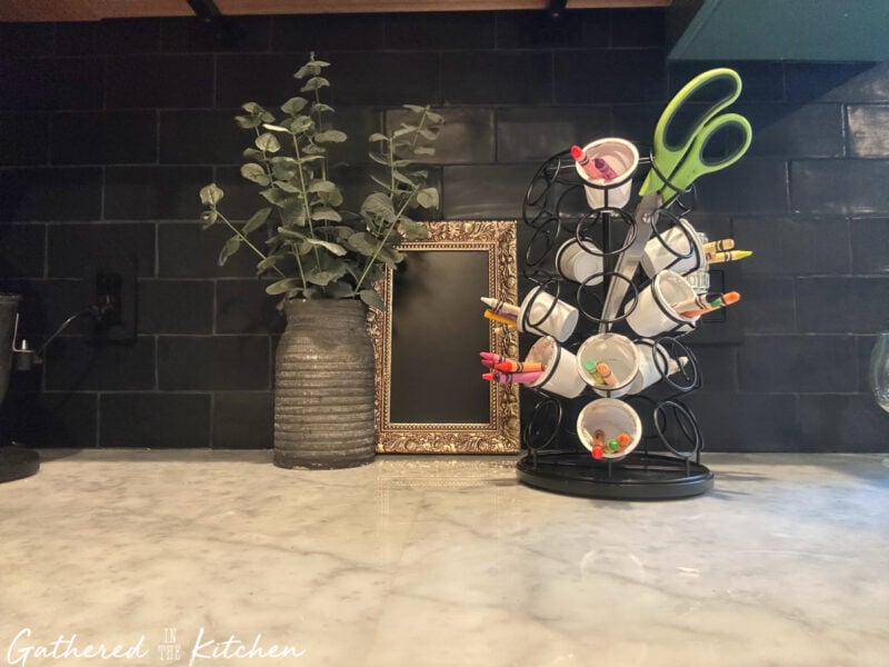 Repurposed Keurig coffee pod holder used as a craft supply organizer, filled with markers, crayons, and scissors, sitting on a marble countertop next to a decorative vase and gold frame. | Gathered In The Kitchen 