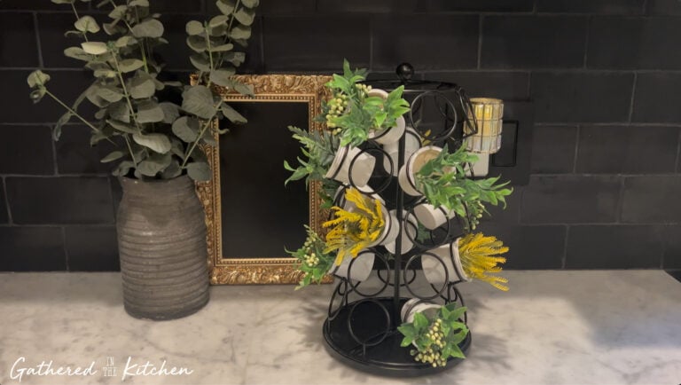 A repurposed Keurig K-Cup holder turned into a decorative faux plant display with greenery and yellow florals, sitting on a marble kitchen counter beside a vintage frame and vase. | Gathered In The Kitchen