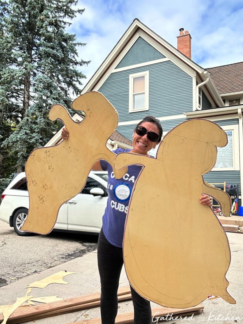 Two large plywood ghost cutouts held up in front of a historic home, ready to be painted and displayed as DIY outdoor Halloween yard decorations. | Gathered In The Kitchen 