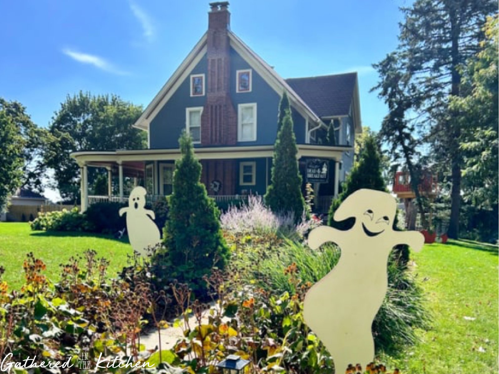 Front yard of a historic blue Victorian-style home decorated for Halloween with two smiling plywood ghost cutouts standing in the garden as friendly outdoor yard decorations. | Gathered In The Kitchen