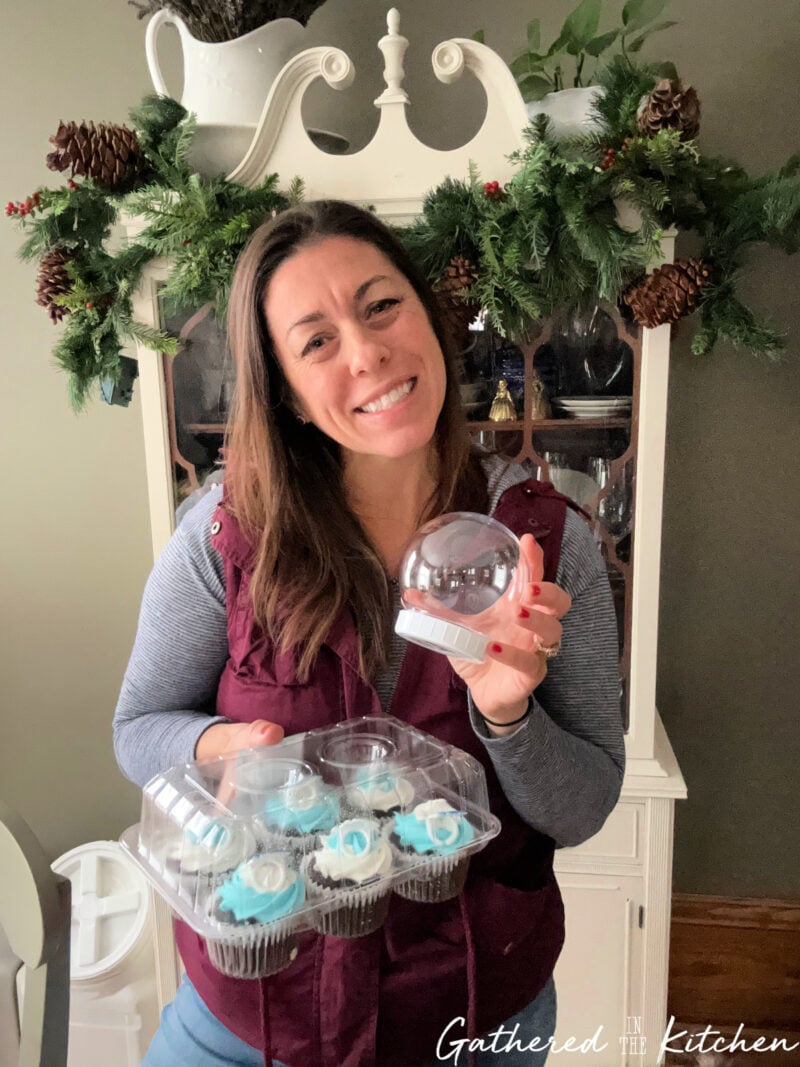 Stephanie smiling and holding a clear plastic snow globe and a container of snowman-themed cupcakes, showing her easy Dollar Tree snow globe cupcake project for the holidays. | Gathered In The Kitchen 