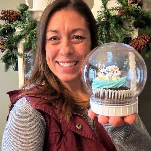Stephanie smiling and holding a finished Dollar Tree snow globe cupcake with a snowman-themed cupcake sealed inside a clear plastic dome — an easy DIY Christmas dessert idea. | Gathered In The Kitchen