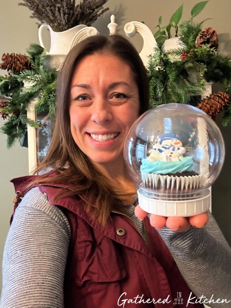 Stephanie smiling and holding a finished Dollar Tree snow globe cupcake with a snowman-themed cupcake sealed inside a clear plastic dome — an easy DIY Christmas dessert idea. | Gathered In The Kitchen 