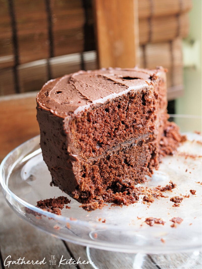 A slice of homemade chocolate cake with rich chocolate frosting on a glass cake stand, showing its moist texture made with boiling water for extra flavor and softness. | Gathered In The Kitchen