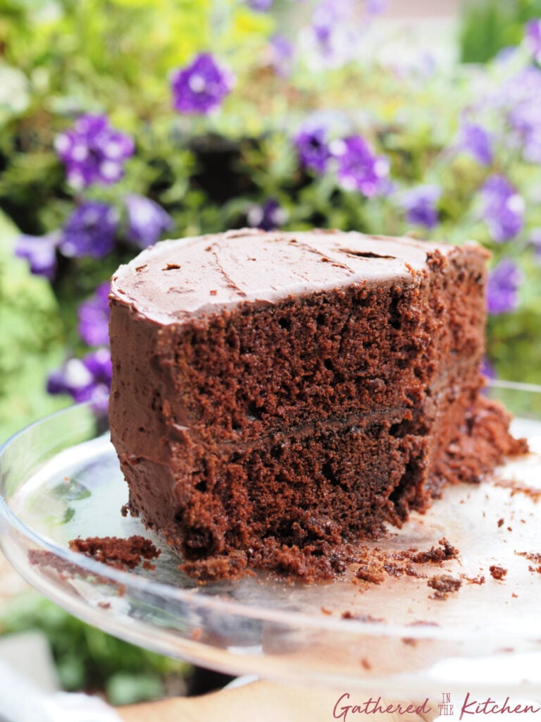 A slice of homemade chocolate cake with rich chocolate frosting on a glass cake stand, showing its moist texture made with boiling water for extra flavor and softness. | Gathered In The Kitchen