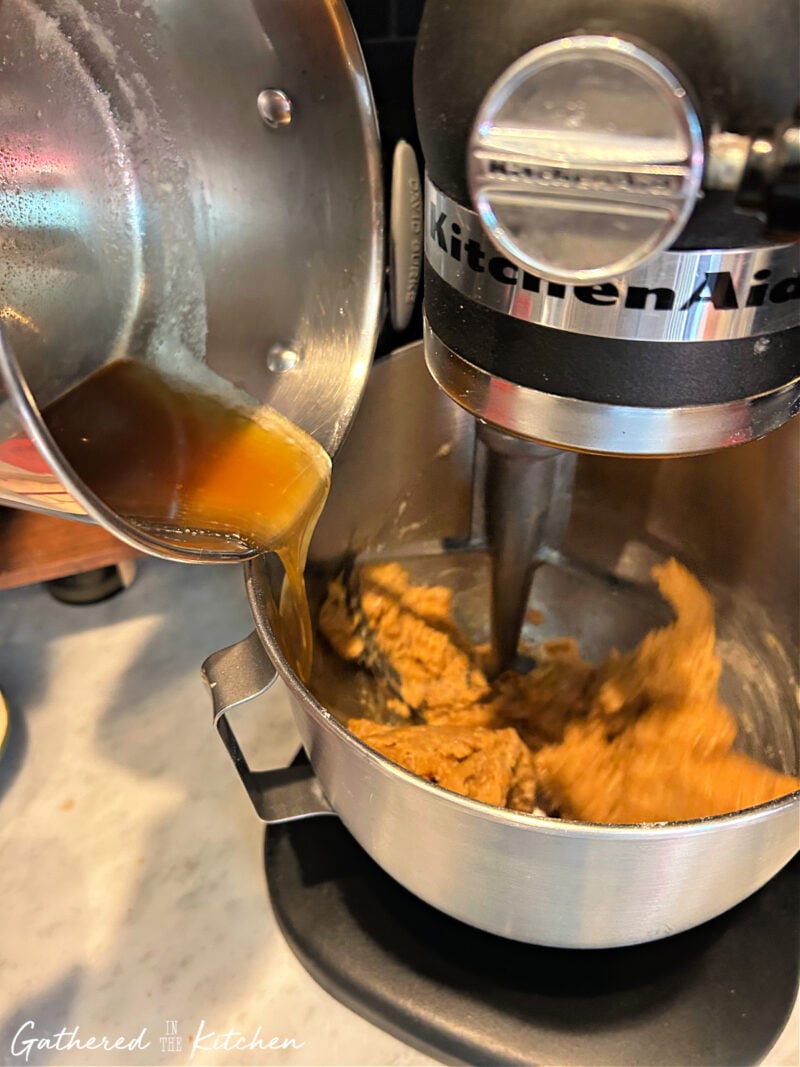 A saucepan of warm browned butter being poured into a mixing bowl with pumpkin cookie dough in a KitchenAid mixer, showing the key step for making chewy pumpkin chocolate chip cookies. | Gathered In The Kitchen 