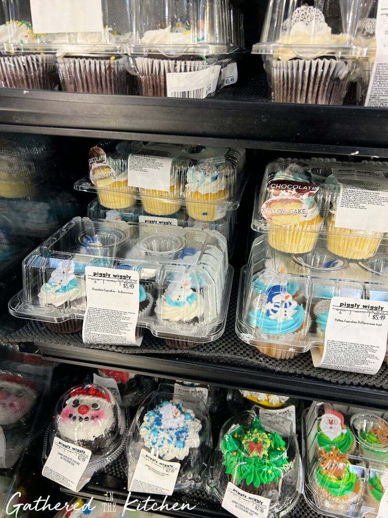 Assorted store-bought holiday cupcakes with snowman and Santa designs displayed in plastic containers at Piggly Wiggly — perfect for making easy Dollar Tree snow globe cupcakes or Christmas party desserts. | Gathered In The Kitchen 