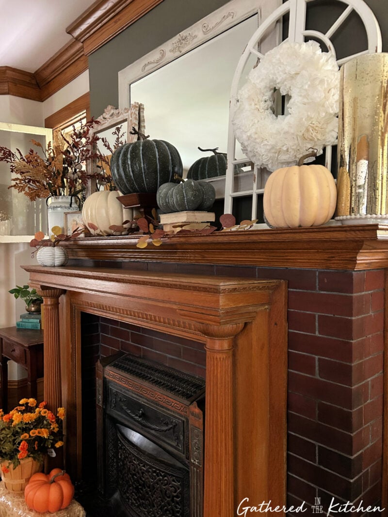 Victorian oak fireplace mantel decorated for fall with green and white pumpkins, a white wreath, vintage books, autumn leaves, and warm seasonal accents in a historic home. | Gathered In The Kitchen 