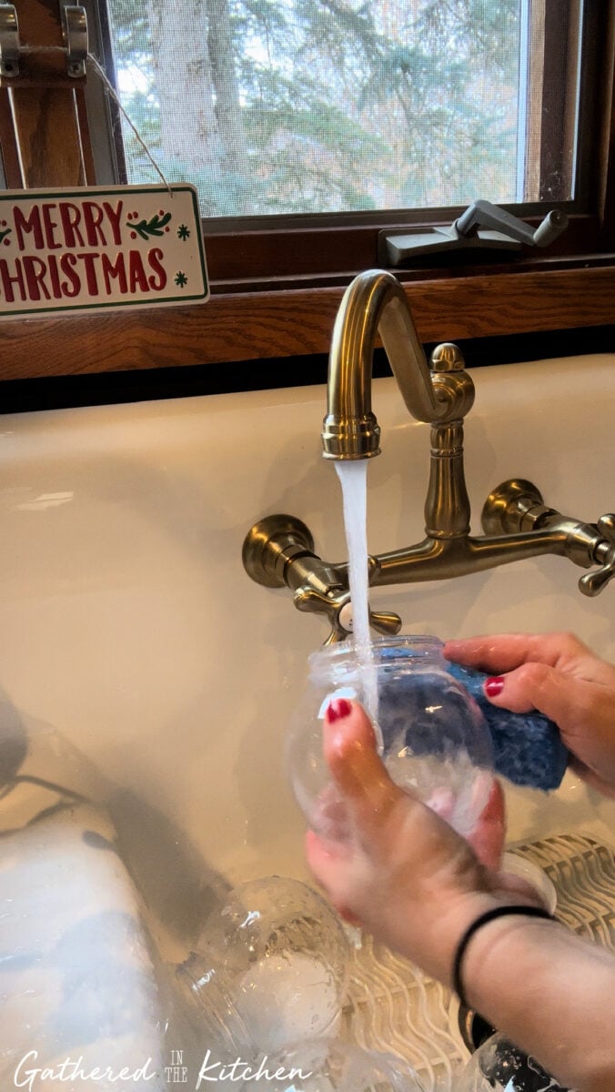Hands washing a clear plastic Dollar Tree snow globe under running water in a farmhouse sink with a “Merry Christmas” sign in the background — prepping for DIY snow globe cupcakes. | Gathered In The Kitchen 