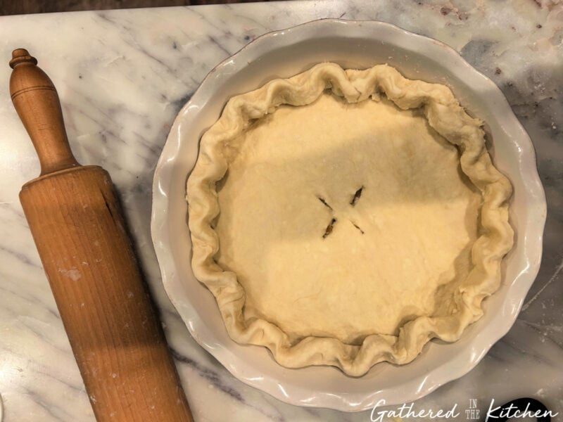 Homemade pie crust in a pie dish with crimped edges and a vent cut in the top, sitting beside a wooden rolling pin on a marble countertop. | Gathered In The Kitchen 
