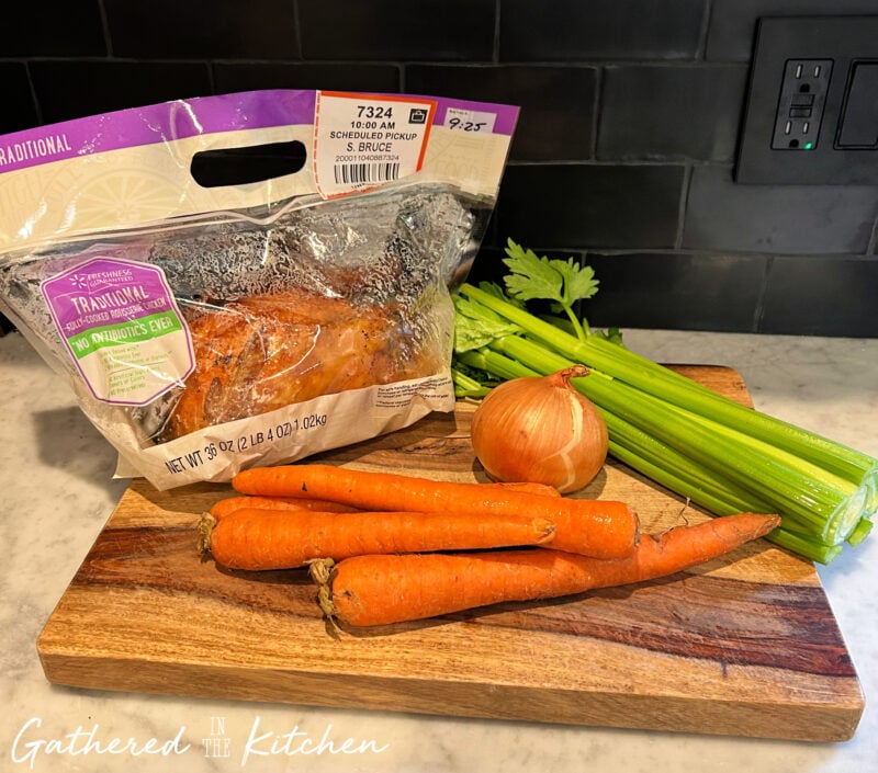 Rotisserie chicken, carrots, celery, and an onion arranged on a wooden cutting board, showing the fresh ingredients used to make chicken pot pie. | Gathered In The Kitchen 