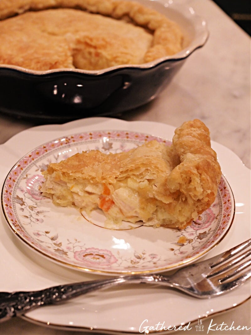 Slice of homemade chicken pot pie on a decorative floral plate, showing the flaky golden crust and creamy rotisserie chicken filling, with the full pie in the background. | Gathered In The Kitchen 