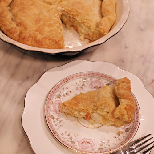 Slice of homemade chicken pot pie on a decorative plate with a fork, with the remaining pie in the background. | Gathered In The Kitchen