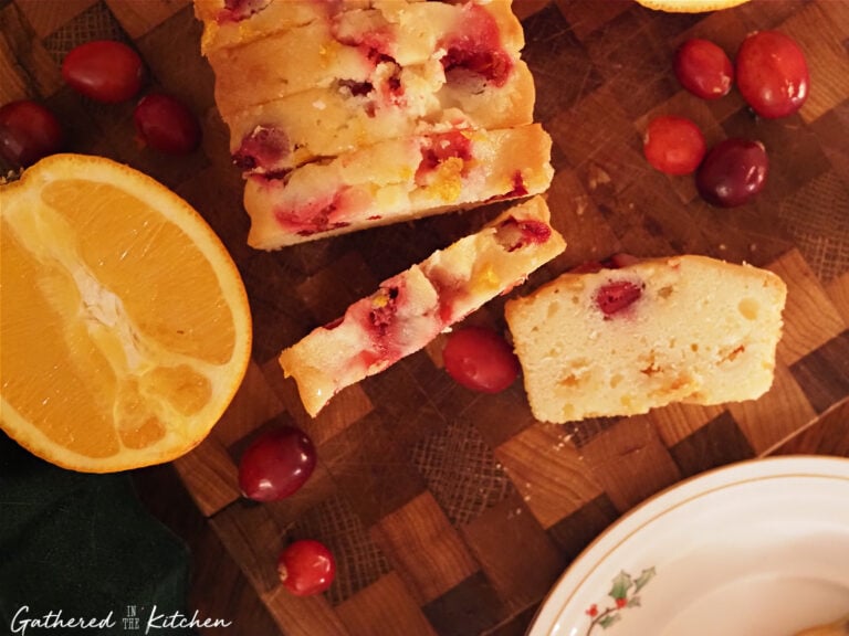 Overhead view of sliced cranberry orange pound cake on a wooden cutting board with fresh cranberries and an orange half, a festive Christmas bread and homemade baked good for neighbor gifts | Gathered In The Kitchen